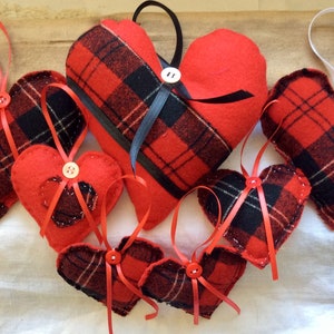 Puede incluir: Siete adornos de corazón de fieltro rojo y negro a cuadros con acentos de cinta blanca y roja. Cada corazón tiene un botón en el centro.