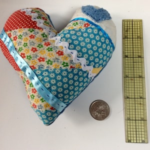 Pode incluir: Uma almofada em forma de coração feita de patchwork com tecido azul, vermelho, branco e amarelo. A almofada tem aproximadamente 15 cm de largura e 13 cm de altura.