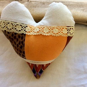 Può includere: Un cuscino a forma di cuore realizzato in tessuto patchwork. Il cuscino è rivestito con una varietà di tessuti, tra cui stampa leopardata, arancione e motivi floreali. Il cuscino è rifinito con pizzo bianco.