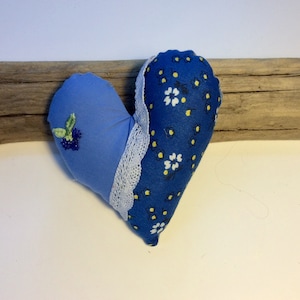 May include: A blue and white heart-shaped pillow with a floral pattern and a small embroidered flower design. The pillow is made of fabric and has a lace trim.