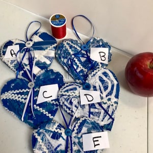 May include: Five blue and white fabric heart ornaments with white ribbon and a button. Each heart has a white paper tag with a letter A through E written on it. A spool of red thread is in the background.
