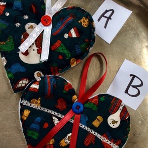 Könnte beinhalten: Zwei Filz-Herzornamente mit winterlichem Muster. Ein Ornament hat ein weißes Band und einen roten Knopf, das andere ein rotes Band und einen blauen Knopf. Die Ornamente sind mit A und B beschriftet.