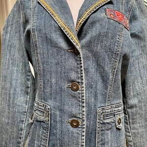 Op de afbeelding: Een blauwe denimjas met gouden bies langs de kraag en zoom. De jas heeft een patch aan de linkerkant met een bloemenmotief. De jas heeft drie knopen aan de voorkant.
