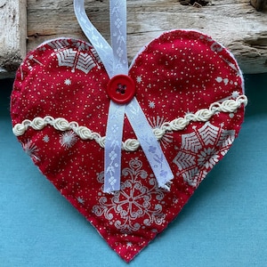 May include: A red fabric heart-shaped ornament with white snowflake designs and a white satin ribbon. The heart has a white lace trim and a red button in the centre.