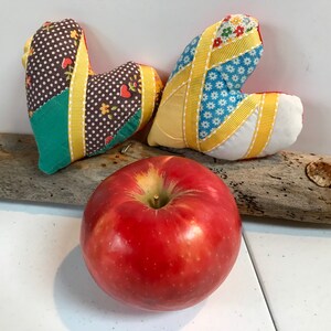 Può includere: Due cuori in tessuto patchwork con accenti di nastro giallo e una mela rossa su un pezzo di legno alla deriva.
