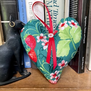 May include: A green heart-shaped cushion with a red ribbon and white flowers and strawberries printed on it. The cushion is sitting on a bookshelf with books behind it.