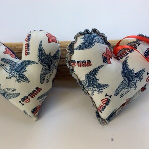 Puede incluir: Dos almohadas de tela blanca en forma de corazón con un estampado azul y rojo de águilas y las palabras "I Love USA". Una almohada tiene una cinta roja.