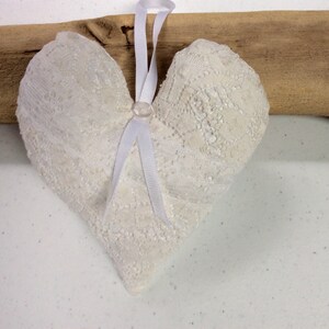 以下が含まれることがあります： 白いレースのハート型のクッション。白いサテンのリボンと白いボタンが付いている。