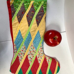 Può includere: Un calzino di Natale in patchwork con un motivo a rombi rosso, verde, giallo e blu. Il calzino è fatto di ritagli di tessuto e ha un polsino in feltro verde. Una mela rossa è appoggiata accanto al calzino.