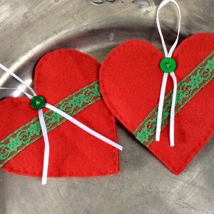 以下が含まれることがあります： 緑のレースのトリムと白いリボンのアクセントが付いた、赤いフェルトのハートのオーナメントが2つ。それぞれのハートの中央には緑のボタンが付いています。