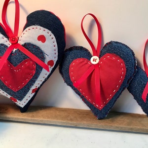 Puede incluir: Tres adornos de corazón de mezclilla con cinta roja y corazones de fieltro rojo. Los corazones son de diferentes tamaños y tienen costuras blancas alrededor de los bordes.