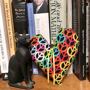 May include: A heart-shaped cushion with a black background and colourful peace signs. The cushion is sitting on a shelf with books behind it.