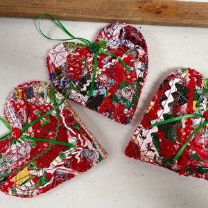 Op de afbeelding: Drie handgemaakte hartvormige kerstversieringen. Elk ornament is gemaakt van rode stof met een patchwork-ontwerp, groen lint en een rode knoop. De ornamenten hebben een groene lintlus om op te hangen.