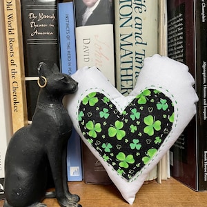 May include: A white heart-shaped pillow with a black and green shamrock pattern on the front. The pillow is sitting on a bookshelf with several books visible.