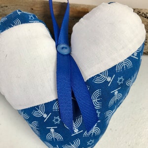 以下が含まれることがあります： メノラー柄の青と白のハート型のクッション。青いリボンとボタンが付いています。クッションの前面には白い布のパネルがあります。