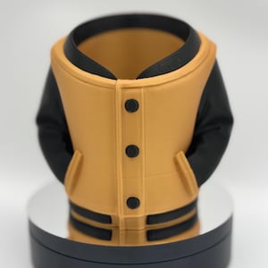 Peut inclure: Objet décoratif imprimé en 3D en forme de veste universitaire. La veste est jaune moutarde avec des manches, un col et des bordures noirs. Elle comporte trois boutons noirs et deux rayures noires en bas.