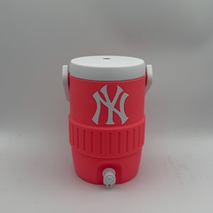 May include: A bright pink beverage cooler featuring a white lid and a white New York Yankees logo on the front. The cylindrical container includes a white handle on each side and a white spigot at the bottom for dispensing liquids.