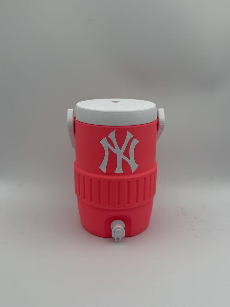 May include: A bright pink beverage cooler featuring a white lid and a white New York Yankees logo on the front. The cylindrical container includes a white handle on each side and a white spigot at the bottom for dispensing liquids.