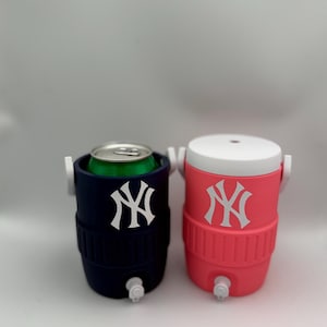 May include: Two small beverage coolers, one navy blue and one pink, each featuring a white New York Yankees logo on the front. The navy cooler holds a green aluminum can, while the pink cooler has a white lid. Both have white spigots at the base.
