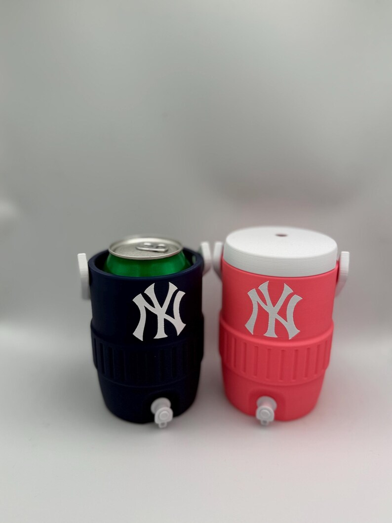 May include: Two small beverage coolers, one navy blue and one pink, each featuring a white New York Yankees logo on the front. The navy cooler holds a green aluminum can, while the pink cooler has a white lid. Both have white spigots at the base.