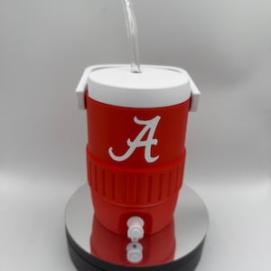 May include: A red and white cooler-style tumbler with a clear straw. The tumbler features a white "A" logo on the front. It has a white lid and handle, and a spigot at the bottom. The tumbler is designed to keep drinks cold.