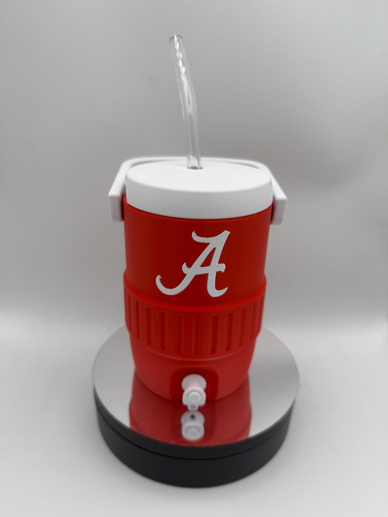May include: A red and white cooler-style tumbler with a clear straw. The tumbler features a white "A" logo on the front. It has a white lid and handle, and a spigot at the bottom. The tumbler is designed to keep drinks cold.