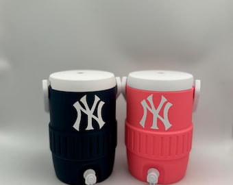 Può includere: Due refrigeratori d'acqua isolati, uno blu navy e uno corallo, ciascuno con coperchio e manico bianchi. Il logo bianco dei New York Yankees è ben visibile sulla parte anteriore di ogni refrigeratore. Un rubinetto bianco si trova in basso.