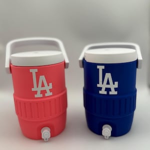 May include: Two insulated water coolers, one coral and one blue, each with a white lid and handle. The coolers feature the white "LA" logo. Each cooler has a white spigot near the bottom. Perfect for keeping drinks cold.