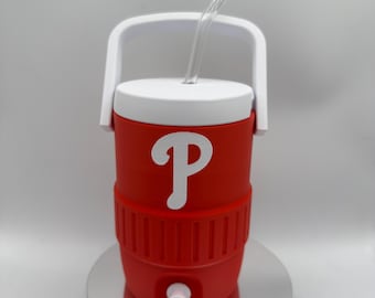 Può includere: Un refrigeratore rosso e bianco con manico e coperchio bianchi. Una cannuccia trasparente si estende dalla parte superiore. Il refrigeratore presenta un logo "P" bianco sul davanti e un rubinetto bianco in basso. Il refrigeratore è su una piattaforma argentata.