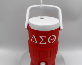 Può includere: Contenitore termico isolato rosso e bianco con manico e coperchio bianchi. Il contenitore ha una cannuccia trasparente e le lettere greche Delta, Sigma e Theta in bianco. Ha un rubinetto bianco.