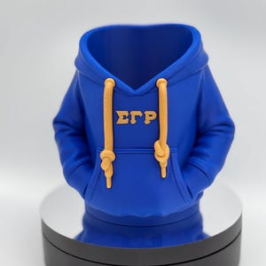 以下が含まれることがあります： フードとポケットが付いた、青いパーカー型の装飾品。 パーカーには黄色のドローストリングと、前面に黄色のギリシャ文字ΣΓΡが付いています。 黒とシルバーの回転スタンドに表示されています。
