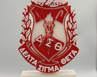 Può includere: Un emblema rosso e bianco con lettere greche e un disegno a torcia, montato su una base rettangolare bianca. Il design include una forma a scudo, foglie di alloro e una figura in alto. Il design è dettagliato.