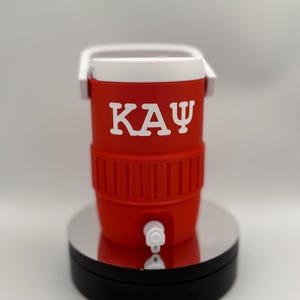 May include: A red and white cooler with the Greek letters K A Ψ in white. The cooler has a white handle and a spigot. The cooler is sitting on a silver and black stand.