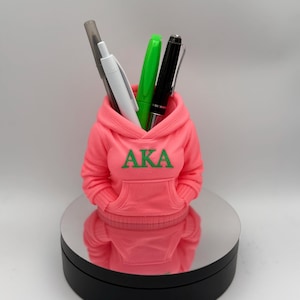 Puede incluir: Un portalápices rosa con forma de sudadera con capucha, con las letras "AKA" en verde. Contiene un bolígrafo gris, un bolígrafo blanco, un bolígrafo verde y un bolígrafo negro. El portalápices está sobre una base giratoria plateada y negra.