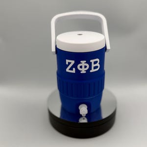 May include: A blue beverage cooler with a white lid, a white handle, and a white spigot at the base. The front of the cooler features the white Greek letters ZΦB printed in a bold, sans-serif font. The cooler has a ribbed texture around the middle.