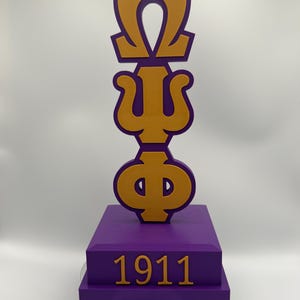 May include: A purple and gold sculpture featuring the Greek letters Omega, Psi, and Phi, on a purple base with the number 1911 in gold. The sculpture is on a black rotating base.