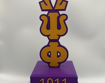 Può includere: Una scultura decorativa viola e oro con le lettere greche Omega, Psi e Phi, su una base viola con il numero 1911 in oro. La scultura è su una base rotante nera.