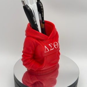 K&ouml;nnte beinhalten: Ein roter Stifthalter in Form eines Hoodies mit wei&szlig;en griechischen Buchstaben. Der Stifthalter enth&auml;lt einen schwarzen Stift, zwei wei&szlig;e Stifte und einen schwarz-silbernen Stift. Der Stifthalter steht auf einer reflektierenden Oberfl&auml;che.