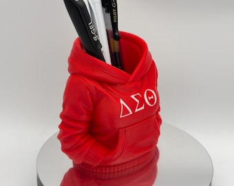 Può includere: Un portapenne rosso a forma di felpa con cappuccio, con lettere greche bianche. Il portapenne contiene una penna nera, due penne bianche e una penna nera e argento. Il portapenne è su una superficie riflettente.