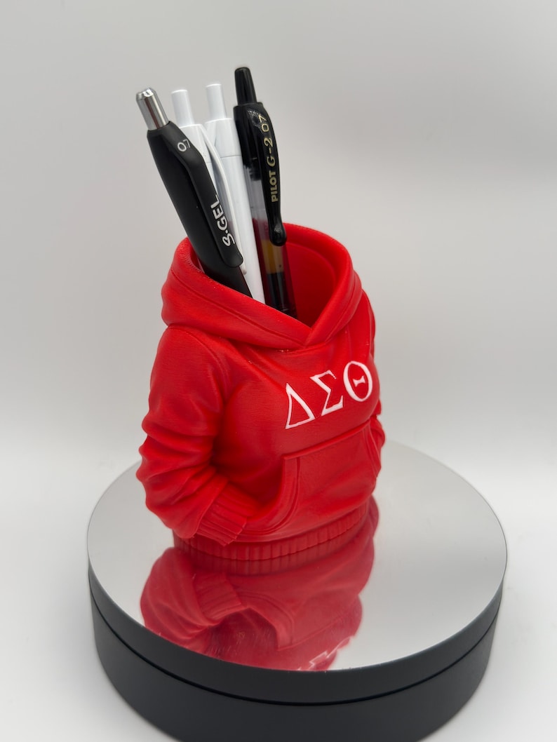 K&ouml;nnte beinhalten: Ein roter Stifthalter in Form eines Hoodies mit wei&szlig;en griechischen Buchstaben. Der Stifthalter enth&auml;lt einen schwarzen Stift, zwei wei&szlig;e Stifte und einen schwarz-silbernen Stift. Der Stifthalter steht auf einer reflektierenden Oberfl&auml;che.