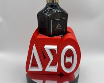Può includere: Una bottiglia nera di sapone per le mani con una pompa bianca è appoggiata su un supporto decorativo rosso e bianco. Il supporto presenta le lettere greche Delta, Sigma e Theta. Il sapone è etichettato 'Mahogany Woods'.