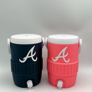 May include: Two insulated water coolers, one navy blue and one coral, each with a white lid and a white "A" logo. The coolers have a spigot at the bottom and white handles. The coolers are designed to keep beverages cold.