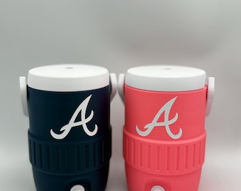 Può includere: Due refrigeratori d'acqua isolati, uno blu navy e uno corallo, ciascuno con un coperchio bianco e un logo "A" bianco. I refrigeratori hanno un rubinetto sul fondo e maniglie bianche. I refrigeratori sono progettati per mantenere le bevande fredde.
