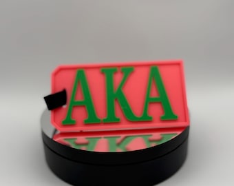 Può includere: Un'etichetta rettangolare rossa con le lettere verdi "AKA". L'etichetta è posta su una base circolare nera. Le lettere sono in grassetto e ben visibili.