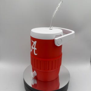 May include: A red and white insulated cooler with a clear straw. The cooler has a white handle and lid, and a white "A" logo. It features a spigot and is designed to keep beverages cold.