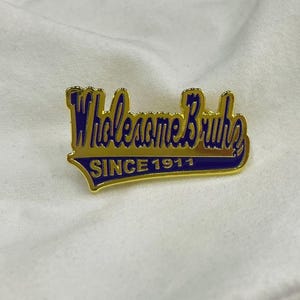 Könnte beinhalten: Goldfarbene Anstecknadel mit dem Schriftzug "Wholesome Bruns" in einem stilisierten Schriftstil und dem Text "SINCE 1911" darunter. Die Nadel hat einen blauen Hintergrund und liegt auf weißem Stoff.