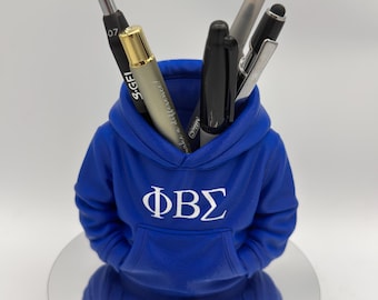 Può includere: Un portapenne blu a forma di felpa con cappuccio, con le lettere greche ΦΒΣ in bianco. Il portapenne contiene diverse penne e uno stilo. Il portapenne è su una superficie riflettente.