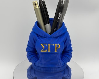 Può includere: Un portapenne blu a forma di felpa con cappuccio, con le lettere greche ΣΓΡ in oro. Il portapenne contiene diverse penne e matite. Il portapenne è su una superficie riflettente.