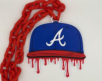 Può includere: Ciondolo a forma di cappellino da baseball blu con logo "A" bianco, bordo rosso e design a goccia di sangue. Il ciondolo è attaccato a una grande catena rossa. Un accessorio unico.