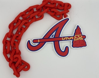 Può includere: Una collana con catena in plastica rossa con un ciondolo raffigurante il logo degli Atlanta Braves. Il ciondolo presenta una "A" rossa e blu con un design a tomahawk. La collana e il ciondolo sono su sfondo bianco.